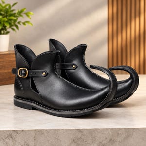 Peut inclure: Une paire de bottines noires dotées d'un bout pointu et recourbé caractéristique. Les bottines présentent une découpe latérale, une fine bride avec une boucle en métal doré, et une semelle plate. La finition noire lisse est soulignée par des coutures.