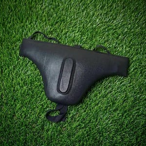 May include: A black protective groin guard featuring a contoured, rigid shell with a central vertical oval reinforcement and integrated elastic straps for secure positioning. The protective gear has a smooth, matte finish and a wide, curved design.