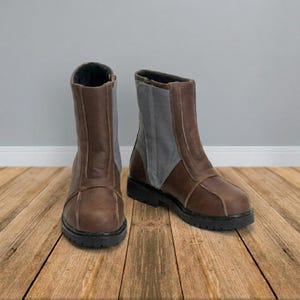 May include: A pair of mid-calf boots featuring brown leather construction with contrasting gray fabric side panels. The boots have a rounded toe, a vertical seam detail along the front, and a thick black lug sole with a defined heel.