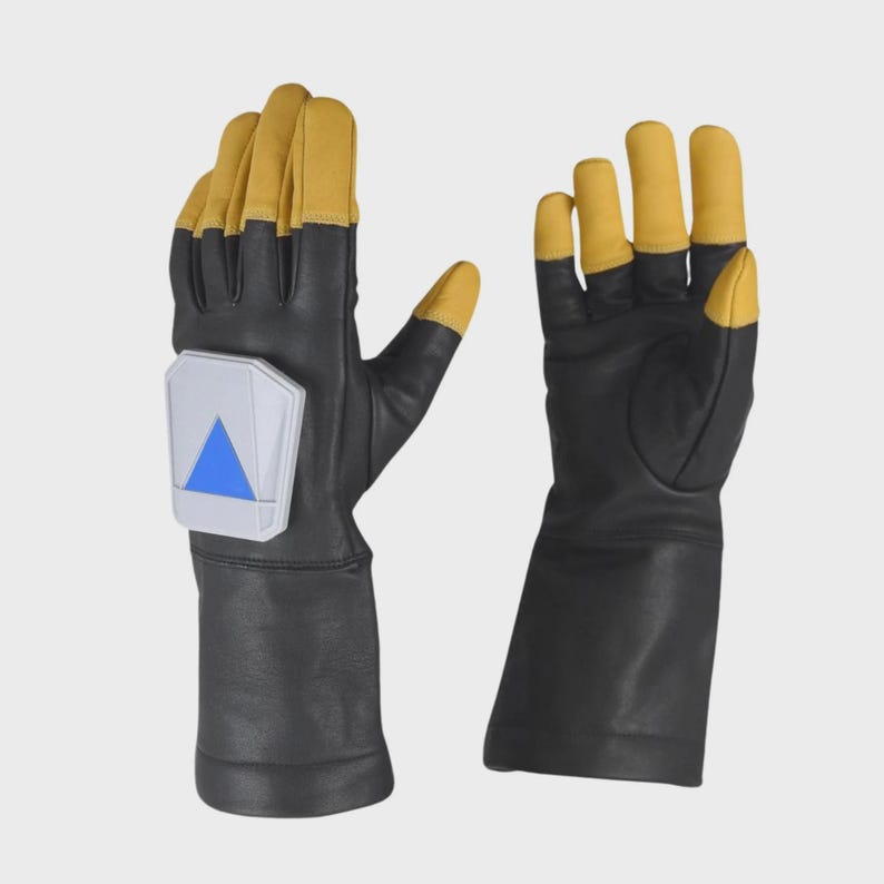 May include: A pair of black and tan gloves with long cuffs. The left glove features a white, pentagonal-shaped rigid plate on the back of the hand, which displays a bright blue triangle pointing downward. The fingers are tipped in tan.