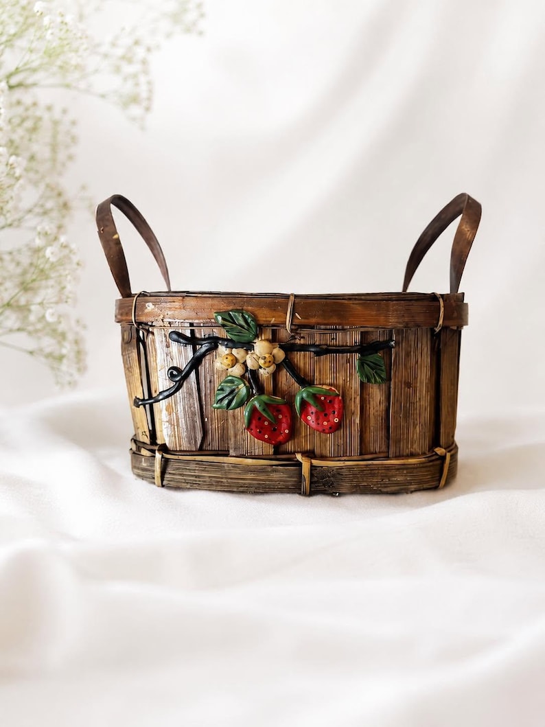 May include: An oval-shaped brown wooden basket featuring two upright handles and a decorative relief of a strawberry vine. The vine includes green leaves, small white flowers, and two red strawberries with visible seeds attached to the front side.