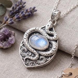 May include: A silver pendant necklace featuring an oval moonstone cabochon at the centre, surrounded by an engraved snake motif with celestial patterns including stars and crescent moons. The pendant hangs from a delicate silver chain.