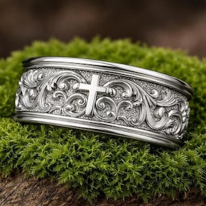 May include: A silver ring with an engraved design. The ring features a cross in the centre and detailed scrollwork. The ring is set against a backdrop of green moss.