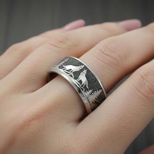 May include: A silver-toned metal ring featuring a textured, dark background with an engraved pattern of two wolves standing among pine trees. The band has a smooth, polished edge and a wide, flat profile that encircles the finger.