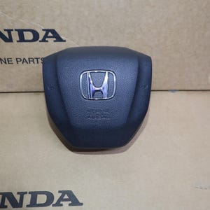 May include: Black Honda SRS airbag cover with the Honda logo in the center. The words "SRS AIRBAG" are printed below the logo. The words "HONDA GENUINE PARTS" are printed on the cardboard background.