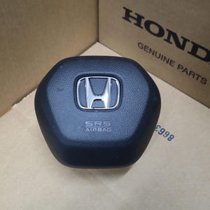 May include: A black Honda steering wheel airbag module with a textured surface, featuring a centered silver Honda emblem and the text SRS AIRBAG embossed below it. The component is positioned in front of a cardboard box labeled HONDA GENUINE PARTS.