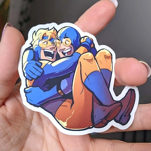 May include: A sticker featuring two cartoon figures embracing. One figure is in blue with a yellow mask, the other in yellow and orange. The sticker is held in a hand.