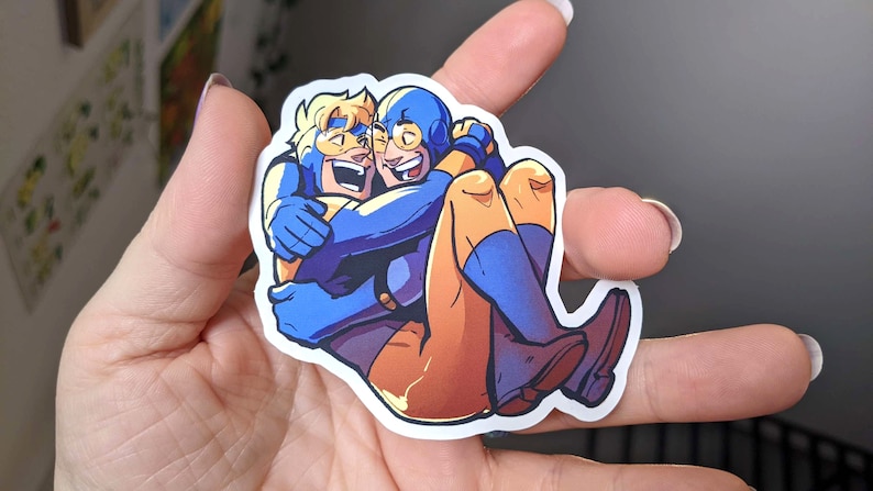 May include: A sticker featuring two cartoon figures embracing. One figure is in blue with a yellow mask, the other in yellow and orange. The sticker is held in a hand.