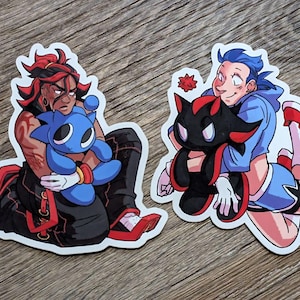 May include: Two die-cut stickers featuring anime-style characters. One sticker depicts a person with red and black hair holding a blue plushie. The other shows a person with blue hair holding a black plushie with red accents.