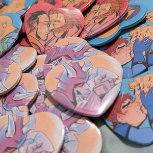 May include: A collection of heart-shaped buttons featuring illustrations of couples in various embraces. The buttons have a glossy finish and are in shades of blue, red, pink, and purple. The illustrations depict romantic scenes.