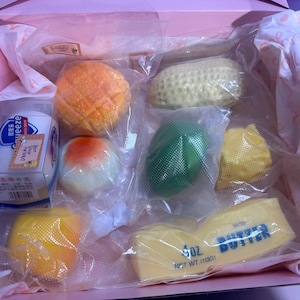 May include: A collection of food-shaped squishy toys inside a pink box, including a melon bun, a peanut, a fried egg, a green fruit, a yellow citrus slice, a yellow fruit, and a block of butter labeled 4 oz. net wt. (113g) in clear packaging.
