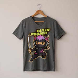 Könnte beinhalten: Ein anthrazitfarbenes V-Ausschnitt-T-Shirt mit einer Grafik einer Ninja-Cartoon-Kreatur mit rosa Haaren und Hörnern, die ein Schwert hält. Über der Figur steht der Text "NINJA POWER" in lindgrünen Buchstaben auf einem dunkelbraunen rechteckigen Hintergrund.