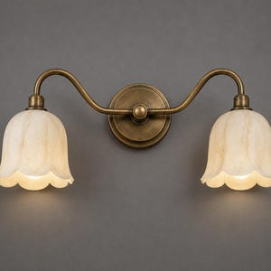 May include: A double wall light fitting featuring a central circular antique brass mounting plate with two curved arms extending outwards, each supporting a bell-shaped, cream-coloured, marbled glass shade with a scalloped edge design.