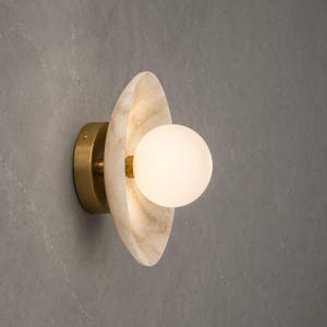 May include: A wall sconce featuring a circular, translucent, cream-coloured stone shade with natural veining. A central, spherical, frosted white glass bulb is mounted on a brass-toned metal base, creating a warm, diffused glow from the centre.