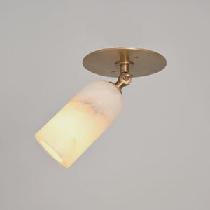 May include: A ceiling-mounted spotlight featuring a circular brass base and a cylindrical, translucent white stone shade. The fixture includes an adjustable brass swivel joint that connects the shade to the mounting plate, allowing for directional light.