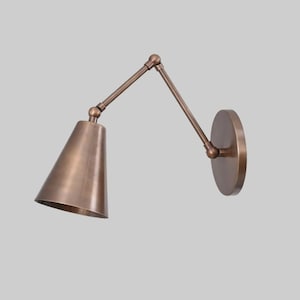 May include: A wall-mounted lamp with a metallic bronze finish, featuring a conical shade and an adjustable articulated arm connected to a circular wall plate. The fixture has a smooth, uniform surface and a functional, multi-jointed design.
