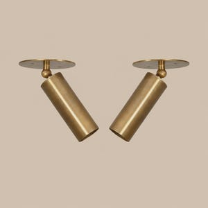 May include: Two matching brass spotlights featuring a cylindrical head attached to a circular mounting plate by a ball-and-socket joint. The metal finish is a muted, brushed gold tone, and each fixture is angled outward to provide directional lighting.