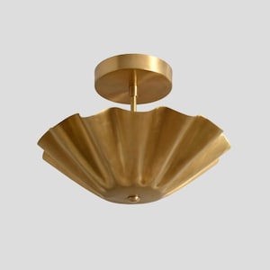 May include: A brass flush mount ceiling light fixture featuring a wide, circular, ruffled shade with a metallic gold finish. The fixture includes a cylindrical central stem connecting the shade to a circular canopy mount at the top.