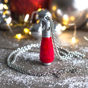 Puede incluir: Un collar de cadena plateada con un colgante de vidrio rojo. El colgante tiene forma de botella y tiene una parte superior plateada. El collar está sobre una superficie de madera con copos blancos como la nieve.