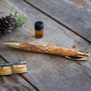 May include: A wooden pen with gold accents, a small brown bottle, and a pair of cufflinks with a wood grain pattern are displayed on a weathered wooden surface. Pine cones and greenery are arranged nearby.