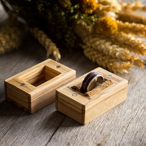 Puede incluir: Dos cajas de madera para anillos con un anillo en una de las cajas. Las cajas son de madera clara con veta natural. El anillo es una banda plateada con una incrustación negra y roja.