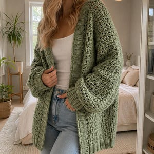May include: An oversized sage green knit cardigan featuring a textured stitch pattern, long sleeves with ribbed cuffs, and a wide ribbed front placket. The garment is worn over a white tank top and light blue denim jeans, creating a layered look.