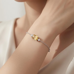May include: A silver-toned chain bracelet featuring a central rectangular yellow gemstone flanked by two small, clear, four-petaled floral-shaped accents on each side. The delicate metal chain secures around the wrist with a simple, refined design.