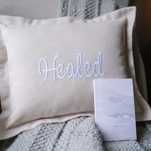 May include: A beige square pillow with the word "Healed" embroidered in white script. A small book with a mountain scene and the text "Healed Everyone" rests beside the pillow on a gray blanket.