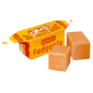 Könnte beinhalten: Zwei quadratische hellbraune Fudge-Bonbons neben einer gelben Verpackung mit einer weißen Kuh-Illustration und dem Text ROSHEN Fudgenta. Die Verpackung hat orangefarbene Wellenlinien und ein spitzes Ende, während die Bonbons eine Textur aufweisen.