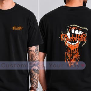 Puede incluir: Una camiseta negra con un pequeño logotipo naranja de Falling in Reverse en el pecho izquierdo. La parte posterior muestra un gráfico grande de una boca con colmillos afilados que gotean líquido naranja, el texto Falling in Reverse y I'm Not A Vampire.