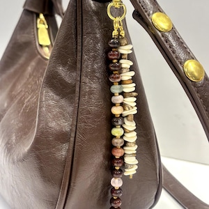 May include: A dark brown leather hobo bag with a gold-tone metal clasp attached to a decorative charm. The charm features two vertical strands: one with multi-coloured round beads and another with flat, off-white, and tan irregular stone discs.
