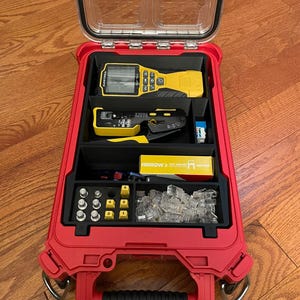 May include: A red, rectangular, hard-shell carrying case with a clear lid, containing various tools and components. Inside are a yellow Klein Tools tester, a black and yellow crimping tool, a box of Arrow staples, and small metal connectors.