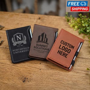 May include: Three rectangular pocket notebooks in black, dark brown, and light brown, each featuring a silver pen held in a side loop. The black notebook displays a crest with the letter N and text reading Northfield University Est. 1867.