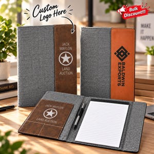 May include: Two gray fabric portfolios with brown leather-like vertical accents. One features the text "JACK WAYLON LAND AUCTION" with a star logo, the other "BALDWIN EXPORTS" with a geometric logo. An open portfolio displays a notepad and pen.