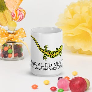 May include: A white ceramic mug featuring a graphic illustration of a marbled newt with a bright yellow and black spotted pattern and a thin orange stripe along its back. The text "MARBLED NEWT" and "TRITURUS MARMORATUS" is printed in black.