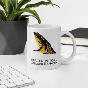 Könnte beinhalten: Ein weißer Keramikbecher mit einer schwarz-gelben grafischen Illustration einer Harlekin-Kröte im Profil. Unter der Kröte steht der Text HARLEQUIN TOAD ATELOPUS HOOGMOEDI in einer schwarzen, handgezeichneten Schriftart. Der Becher hat einen Henkel.