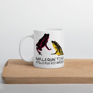 Könnte beinhalten: Ein weißer Keramikbecher mit einer grafischen Illustration von zwei Fröschen. Ein Frosch ist schwarz mit magentafarbenen Mustern, der andere schwarz mit gelben Mustern. Darunter steht der Text HARLEQUIN TOAD ATELOPUS HOOGMOEDI.