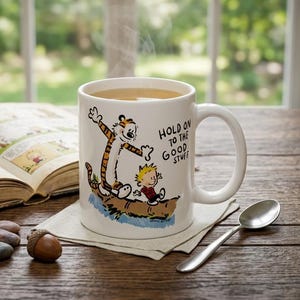 May include: A white ceramic mug filled with a steaming beverage, featuring a graphic of a tiger and a young boy on a log with the text "HOLD ON TO THE GOOD STUFF". The mug sits on a white napkin next to a small silver spoon and an open book.