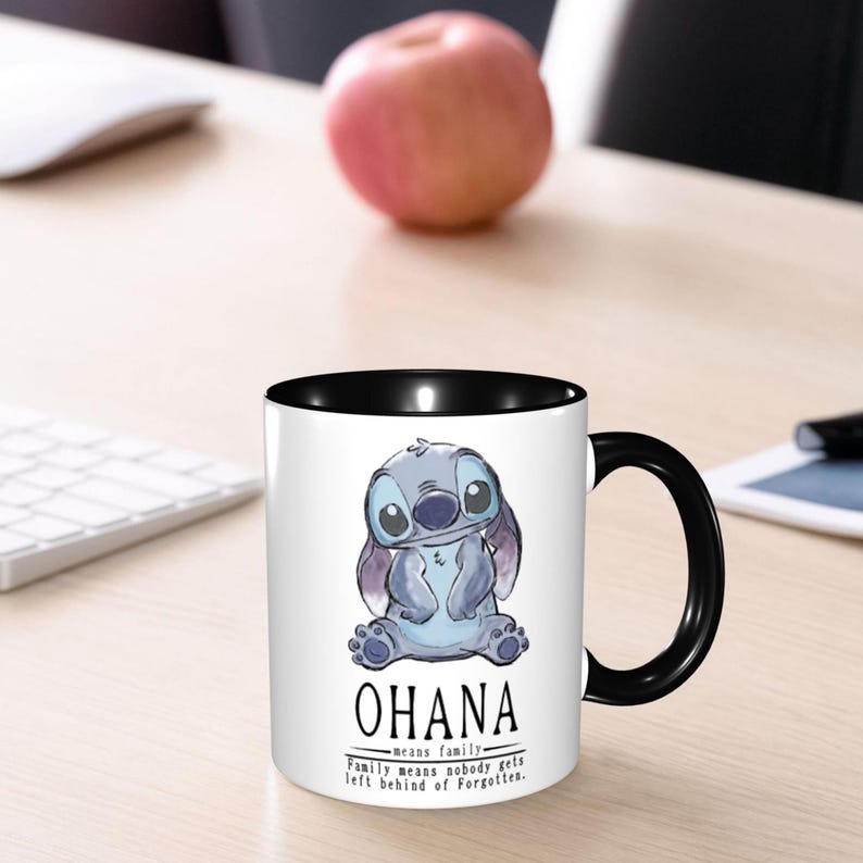 K&ouml;nnte beinhalten: Ein wei&szlig;er Keramikbecher mit schwarzem Innenraum und Henkel, verziert mit einer blauen Alien-Charakter-Illustration und dem Text OHANA means family. Family means nobody gets left behind or forgotten in schwarzer Schrift auf der Vorderseite.