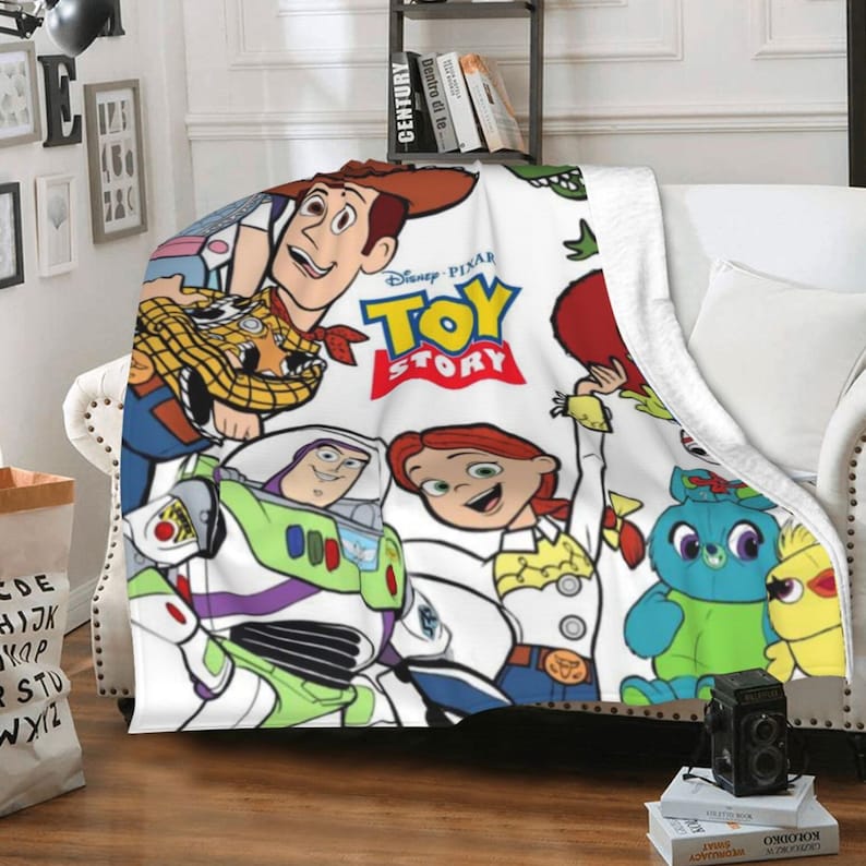 Może przedstawiać: Biały koc z polaru z kolorowym nadrukiem graficznym przedstawiającym postacie z filmu Disney Pixar Toy Story, w tym Chudego, Buzza Astrala, Jessie, Ducky'ego i Bunny'ego. Czerwono-ż&oacute;łte logo Toy Story jest wyraźnie widoczne na przedniej stronie.