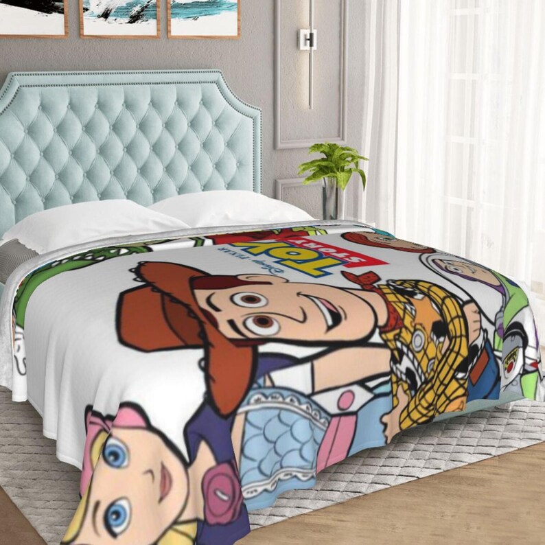 Może przedstawiać: Biały koc z polaru z kolorowym nadrukiem graficznym przedstawiającym postacie z serii Toy Story, w tym Chudego, Buzza Astrala i Pastereczkę, z logo Toy Story wyraźnie widocznym w czerwono-ż&oacute;łtych literach w g&oacute;rnej części.