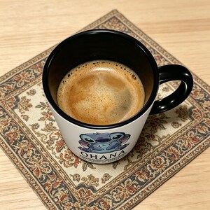 K&ouml;nnte beinhalten: Ein wei&szlig;er Keramikbecher mit schwarzem Innenraum und Henkel, gef&uuml;llt mit dunkelbraunem Kaffee. Die Au&szlig;enseite zeigt eine Grafik einer blauen au&szlig;erirdischen Figur und den Text OHANA, der in schwarzen Gro&szlig;buchstaben unter der Grafik steht.