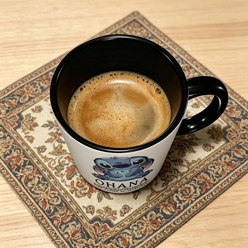 K&ouml;nnte beinhalten: Ein wei&szlig;er Keramikbecher mit schwarzem Innenraum und Henkel, gef&uuml;llt mit dunkelbraunem Kaffee. Die Au&szlig;enseite zeigt eine Grafik einer blauen au&szlig;erirdischen Figur und den Text OHANA, der in schwarzen Gro&szlig;buchstaben unter der Grafik steht.