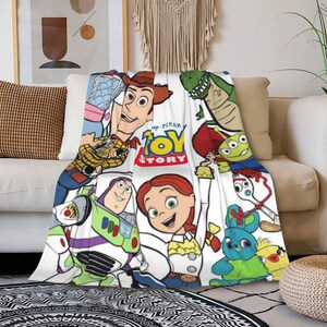 Może przedstawiać: Biały koc z kolorowym nadrukiem graficznym Toy Story. Wz&oacute;r zawiera postacie Chudego, Buzza Astrala, Jessie, Rexa, Sztućka, Kaczora i Kr&oacute;liczka, wraz z logo Toy Story pośrodku, ułożone w powtarzający się wz&oacute;r na całej powierzchni tkaniny.