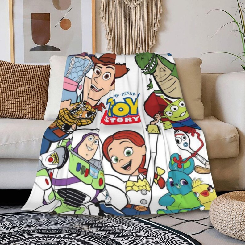 Może przedstawiać: Biały koc z kolorowym nadrukiem graficznym Toy Story. Wz&oacute;r zawiera postacie Chudego, Buzza Astrala, Jessie, Rexa, Sztućka, Kaczora i Kr&oacute;liczka, wraz z logo Toy Story pośrodku, ułożone w powtarzający się wz&oacute;r na całej powierzchni tkaniny.