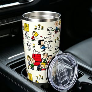 May include: A cream-colored stainless steel tumbler featuring a repeating pattern of Peanuts characters including Snoopy, Charlie Brown, and Lucy. The design includes a red doghouse and a yellow sign that reads 32 COUNT THEM 32. Includes a clear lid.