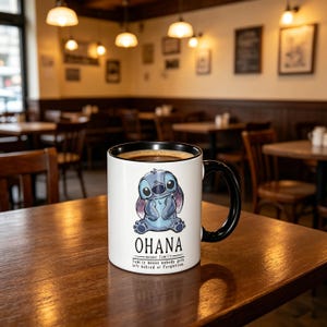K&ouml;nnte beinhalten: Ein wei&szlig;er Keramikbecher mit schwarzem Innenraum und Henkel, der eine blaue Illustration der Figur Stitch zeigt. Unter der Figur steht der Text OHANA means family. Family means nobody gets left behind or forgotten in schwarzer Schrift.