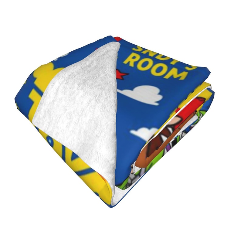 May include: A folded plush blanket featuring a blue background with white clouds and yellow text that reads ANDY'S ROOM. The blanket displays a cartoon character with brown hair and a red hat, with a soft white textured fleece lining visible.