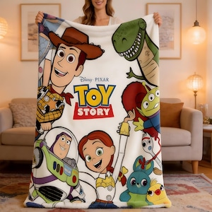 Może przedstawiać: Biały koc z polaru z logo Disney Pixar Toy Story pośrodku, otoczony kolorowymi ilustracjami Chudego, Buzza Astrala, Jessie, Rexa, Obcego, Sztućka, Kaczora i Kr&oacute;lika na miękkiej, teksturowanej powierzchni z tkaniny.