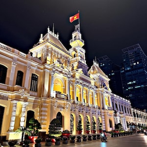 Könnte beinhalten: Das Rathaus von Ho-Chi-Minh-Stadt ist nachts in warmes gelbes Licht getaucht und zeigt eine verzierte französische Kolonialarchitektur mit Rundbogenfenstern und einem hohen zentralen Turm, über dem die rot-gelbe Flagge Vietnams weht.